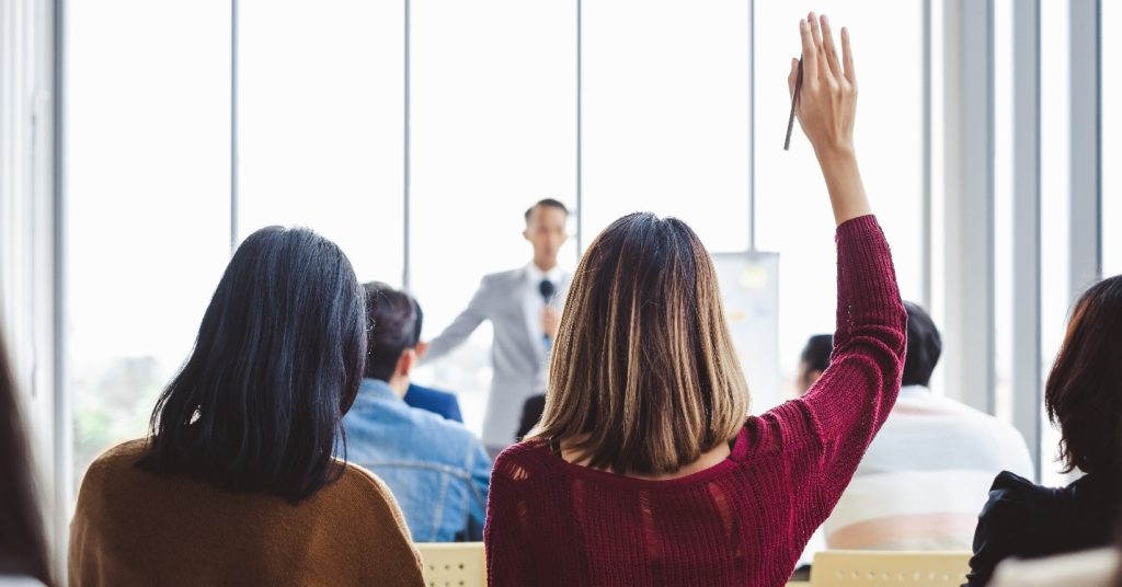People at an event with one woman's hand up to ask a question
